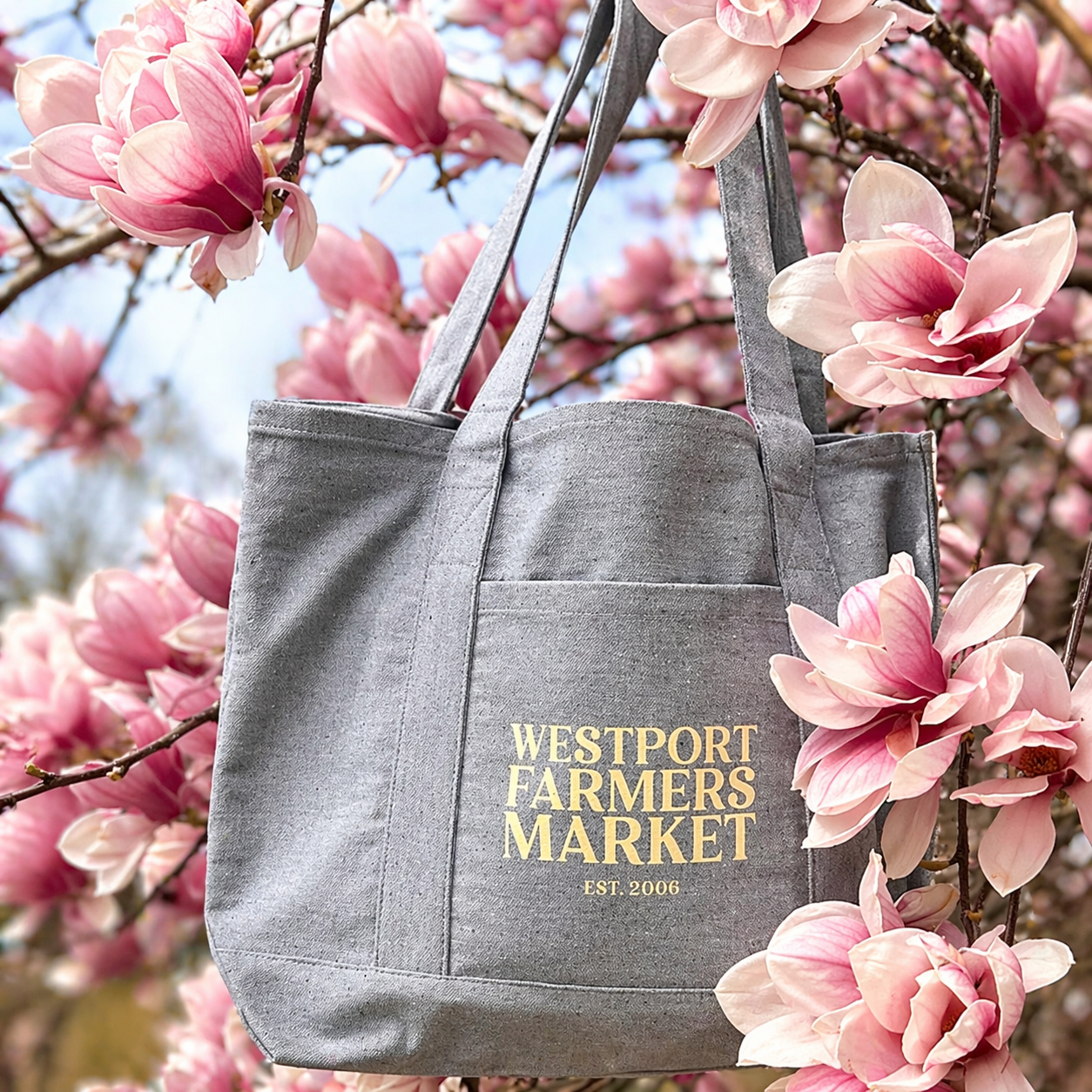 Gray bag with test Westport Farmers Market EST 2006 text on the side. bag is gray. Pink flowers around in background
